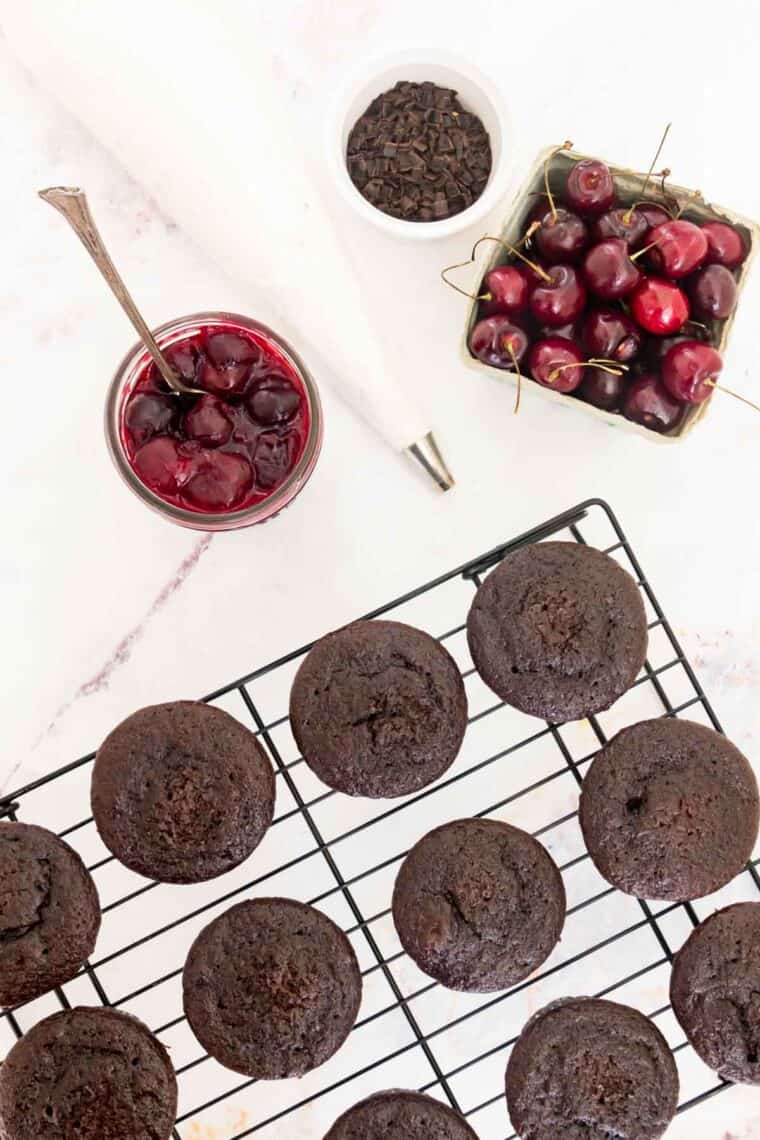 A wire rack holds chocolate cupcakes with a bowl of cherry filling and a box of cherries next to it.