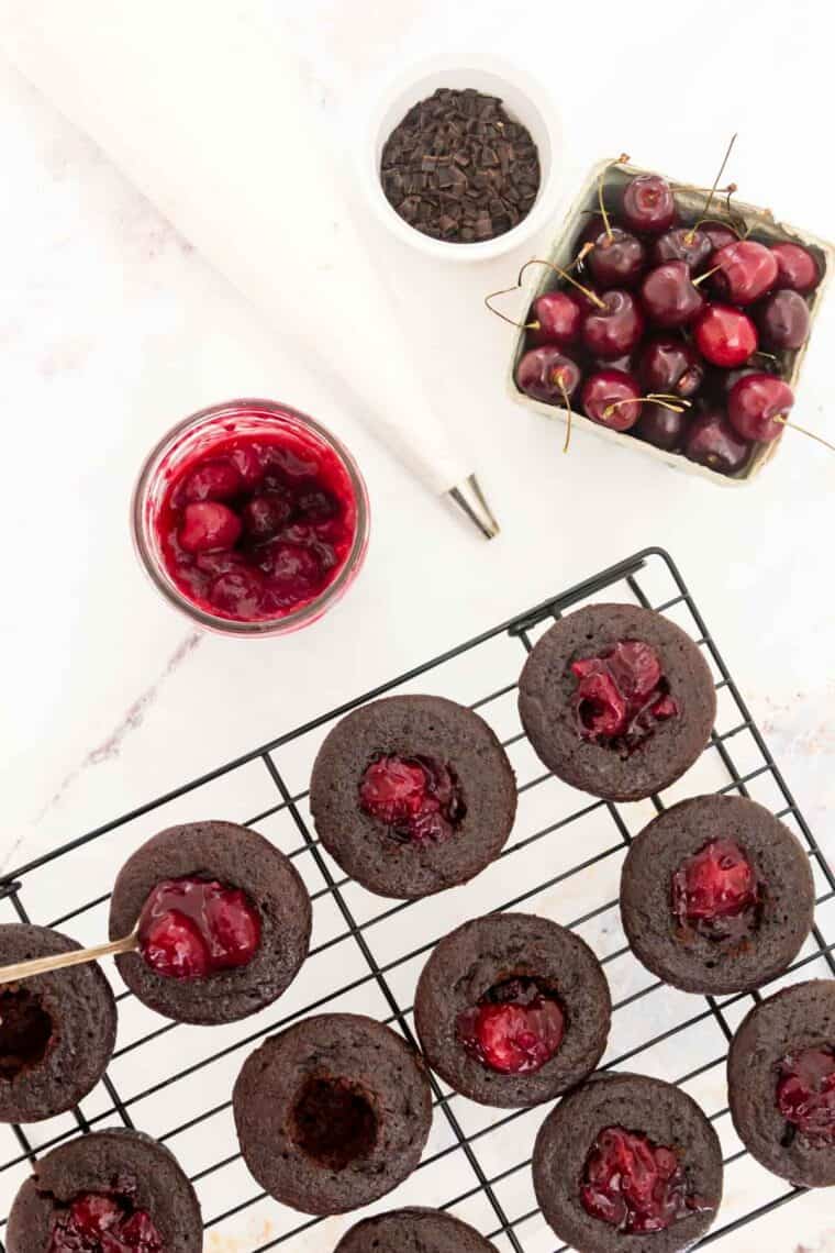 Black forest cupcakes on a wire rack are filled with homemade cherry filling with more cherries nearby.