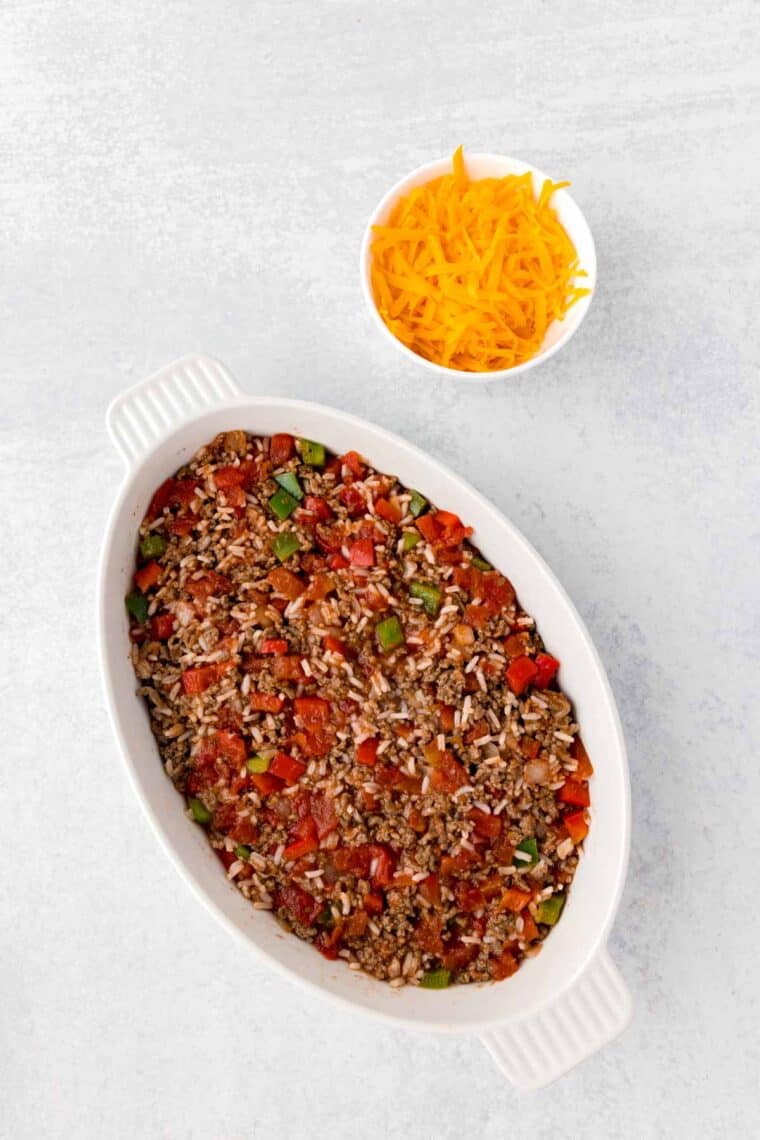 A white casserole dish full of unstuffed pepper casserole with a bowl of shredded cheddar cheese to its right.