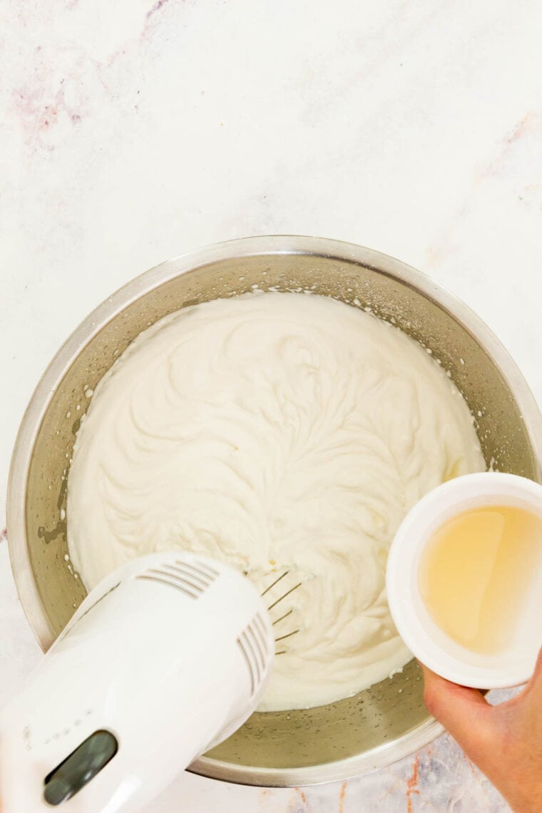 A handheld mixer whips heavy cream while gelatin is poured in.
