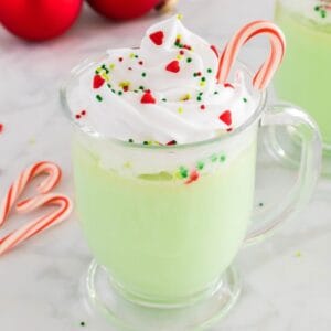 A glass mug of green-colored peppermint white hot chocolate topped with a swirl of whipped cream, festive sprinkles, and a candy cane.