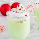 A glass mug of green-colored hot cocoa topped with a swirl of whipped cream, festive sprinkles, and a candy cane with text overlay that says "Peppermint White Hot Chocolate".