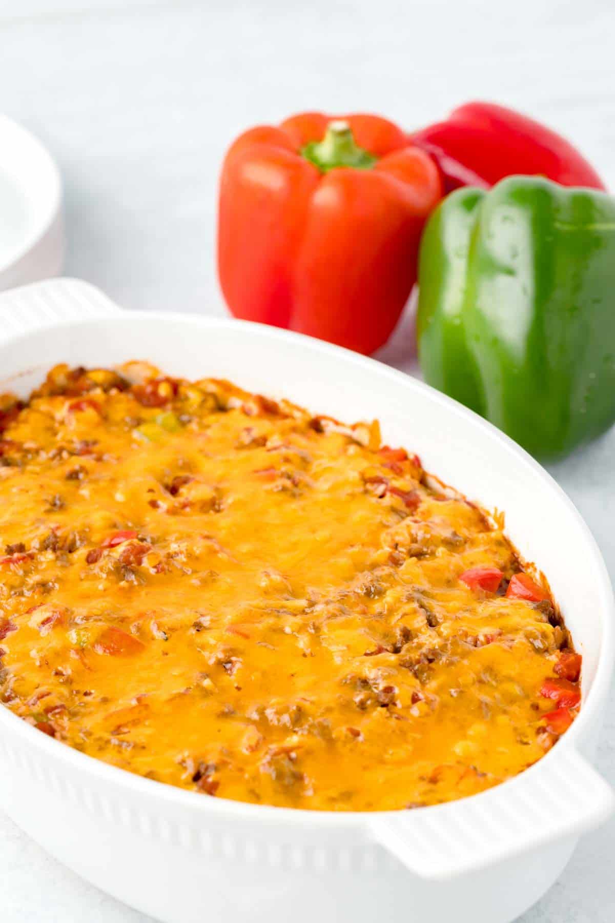 A baked white casserole dish topped with orange melted cheddar cheese with a red and a green bell pepper next to it.