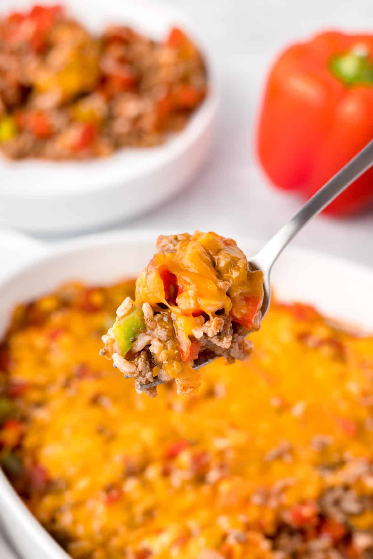 A close up on a spoon holding a bit of unstuffed pepper casserole, with the full casserole seen beneath it and meat and red pepper in the background.