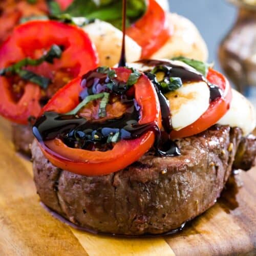 A grilled filet mignon on a wooden board topped with fresh mozzarella and tomato slices and being drizzled with balsamic reduction.