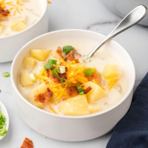 A spoon in a bowl of cream of potato soup topped with bacon, cheddar cheese, and sliced scallions.