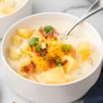 A spoon in a bowl of creamy soup topped with bacon, cheddar cheese, and sliced scallions with text overlay that says "Cream of Potato Soup".