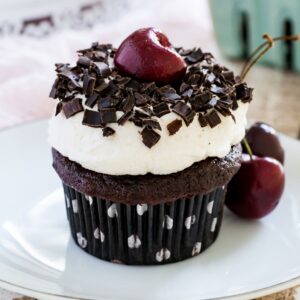 A chocolate cupcake in a polka dot wrapper topped with whipped cream, chocolate shavings, and a cherry.