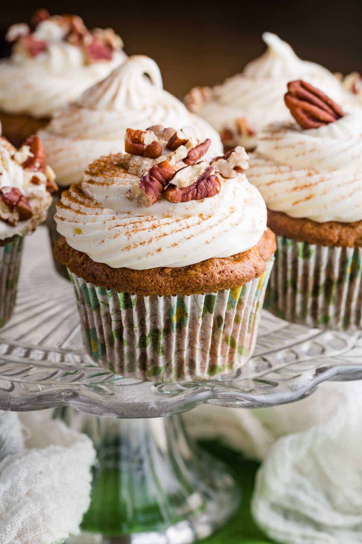 A glass cake stand holds pecan-topped carrot cake cupcakes.