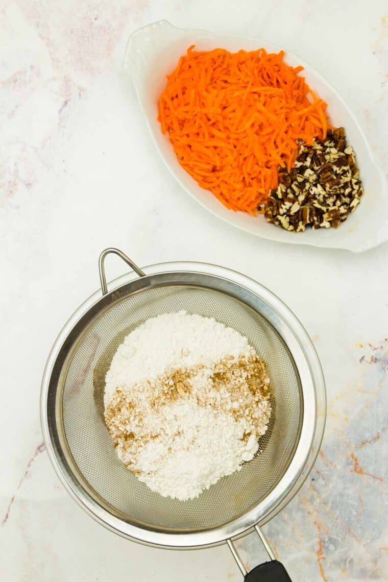 Shredded carrots and pecans in a white dish with a sifter of flour and spices.