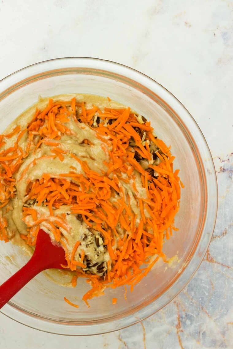 A red spatula stirs shredded carrots and pecans into the batter.