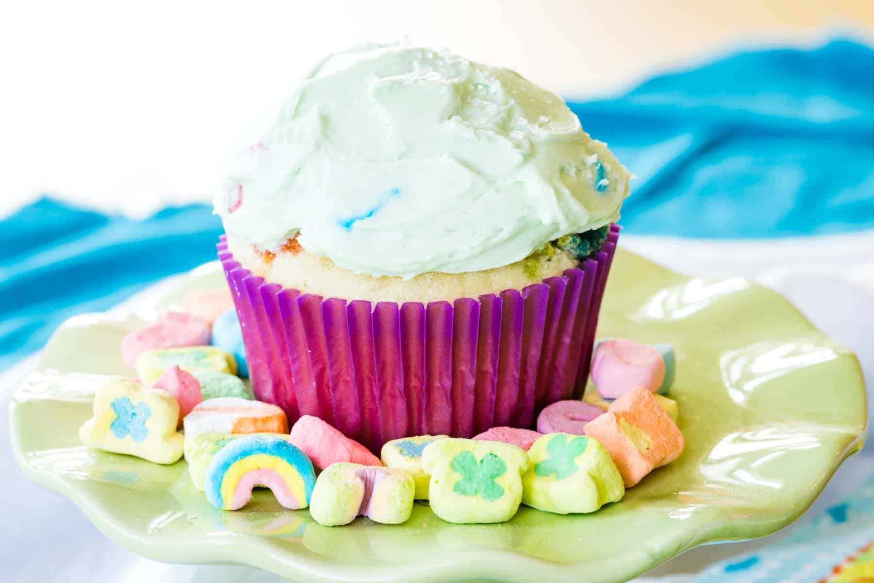 A Lucky Charms cupcake with green frosting is surrounded by Lucky Charms.