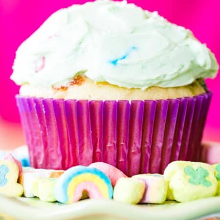 A Lucky Charm cupcake in a pink liner is shown on a cupcake stand surrounded by Lucky Charms.