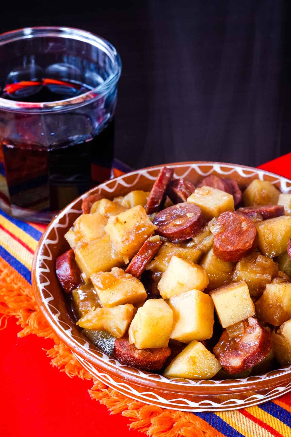 A steaming bowl of patatas riojana is set on a red tablecloth with a glass of red wine in the background.