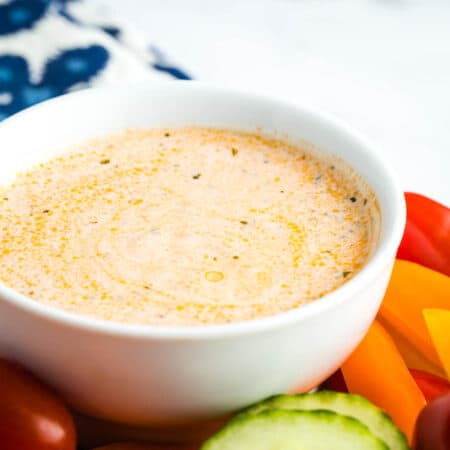 A creamy bowl of buffalo ranch sauce on a table with fresh vegetables alongside it.