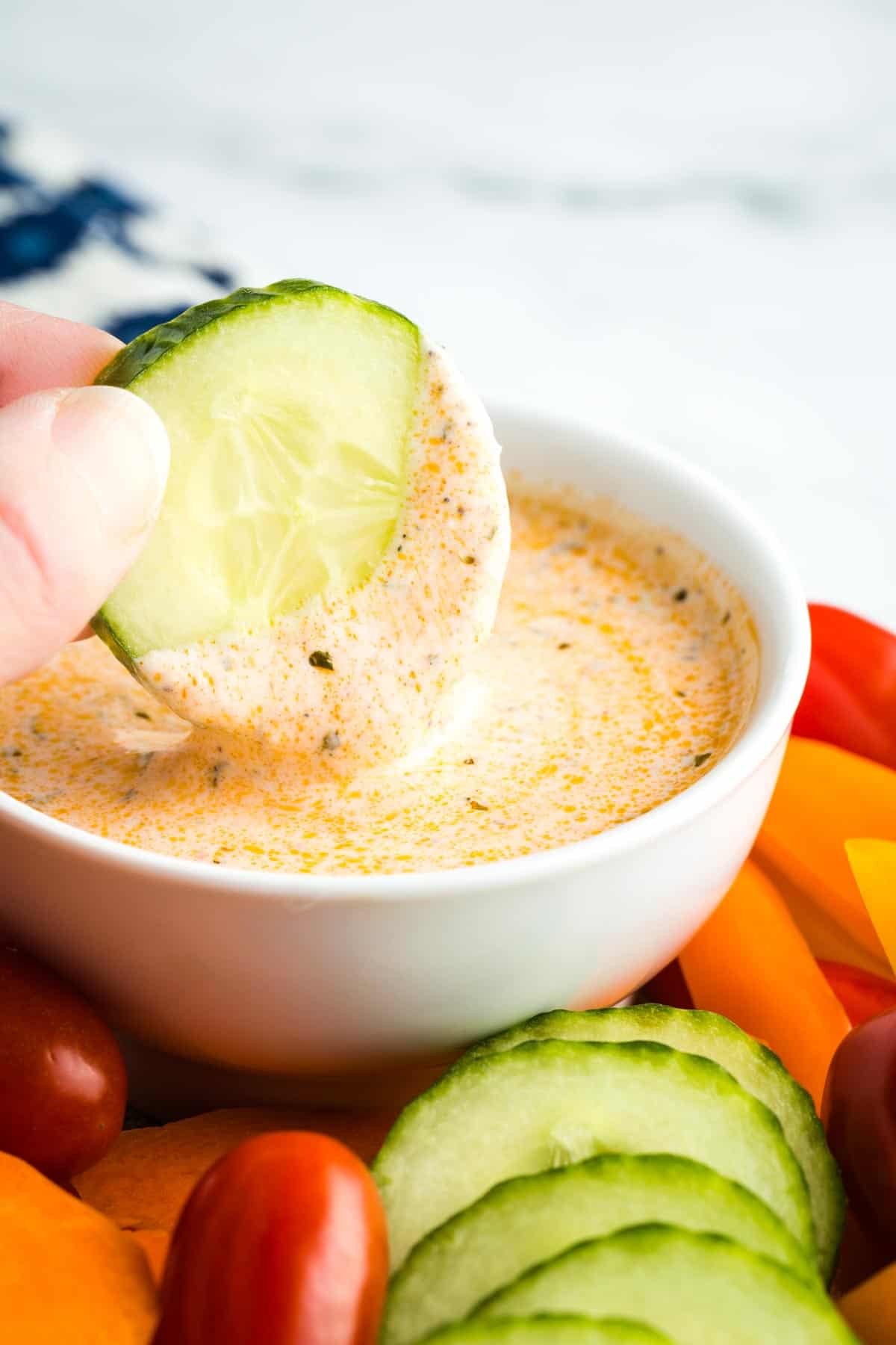 A hand dips a slice of cucumber into a bowl of buffalo ranch sauce.