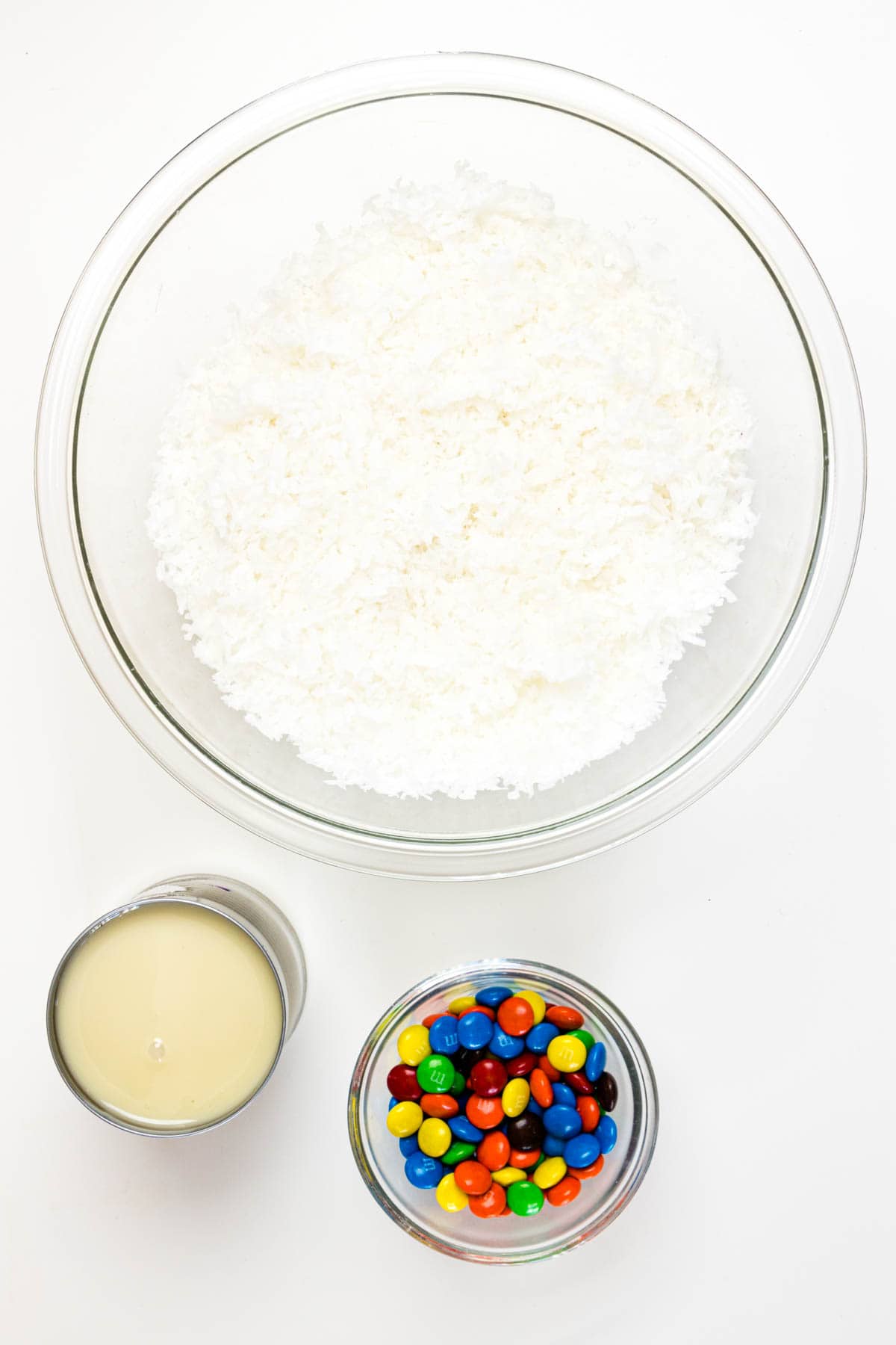 A bowl of shredded coconut, a can of sweetened condensed milk, and a bowl of M&M's.