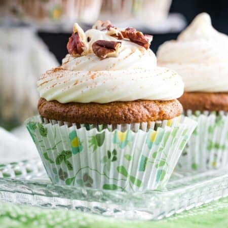 A carrot cake cupcake with cream cheese frosting and chopped pecans on a glass plate.