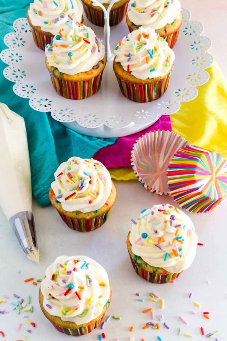 Sprinkles are scattered on and around funfetti cupcakes with a frosting-filled piping bag next to them.