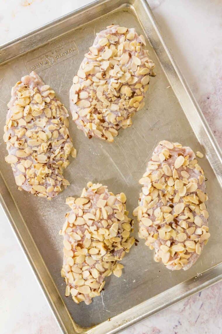 Sliced almonds coat four pieces of chicken on a baking sheet.