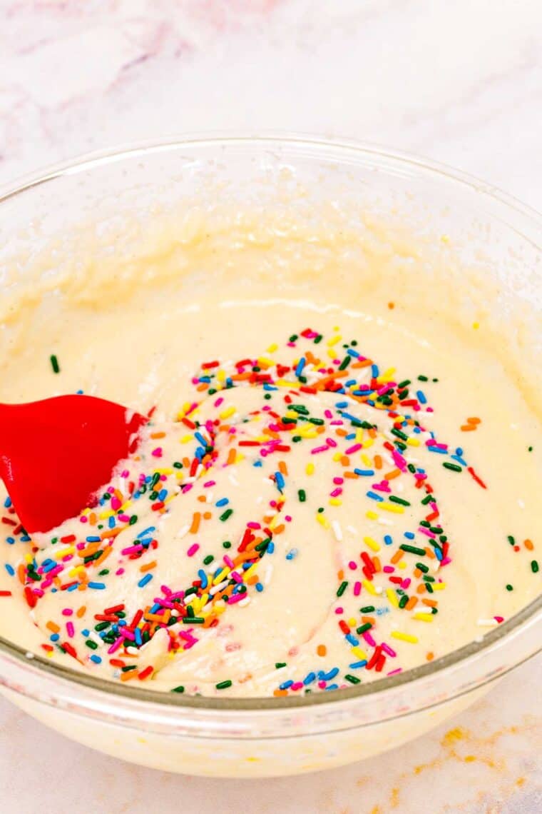 A red spatula gently folds rainbow sprinkles into funfetti cake batter.
