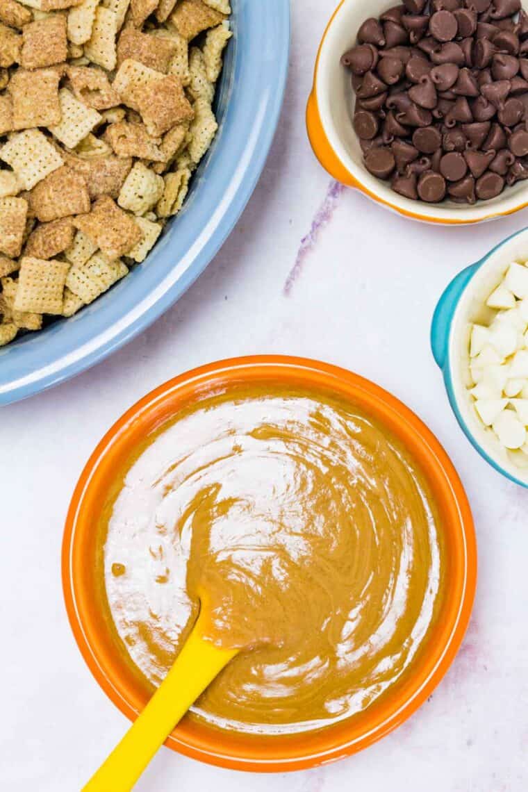 A bowl of peanut butter with a spoon stirring it is next to bowls of cereal, popcorn, and chocolate chips.