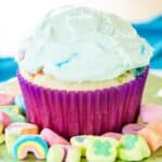 A cupcake with a purple wrapper and green frosting on a platter surrounded by Lucky Charms marshmallows with text overlay that says "Lucky Charms Cupcakes".