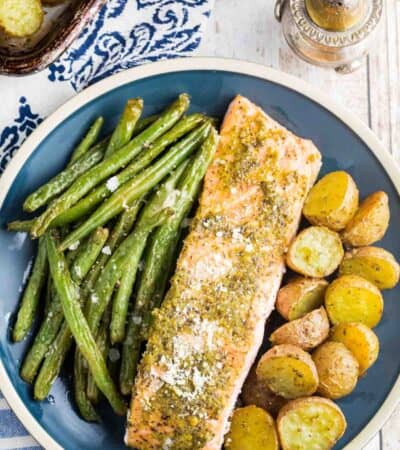A baked piece of pesto salmon on a blue plate with green beans to the left and potatoes to the right.