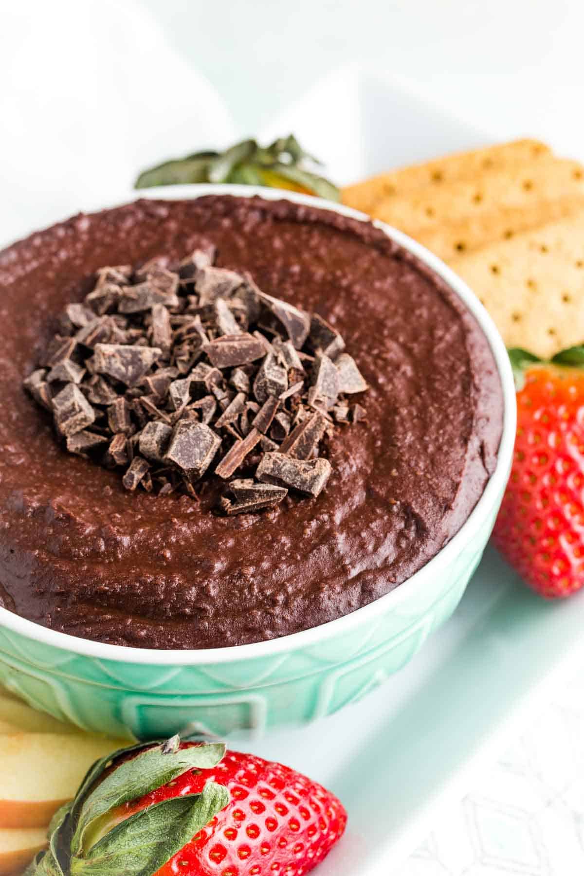 Chopped chocolate on top of a bowl of dark chocolate hummus with fruit and cookies next to it.