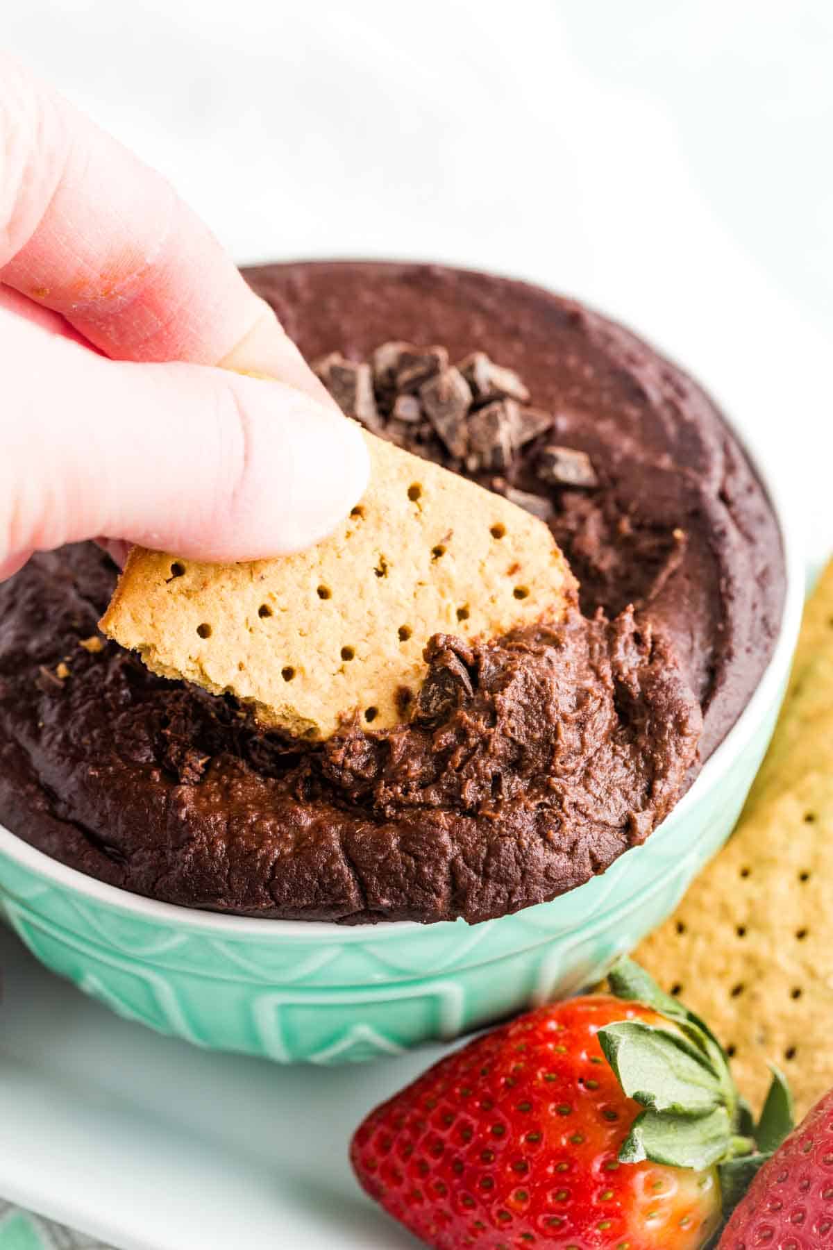 A hand dips a graham cracker into a bowl of dark chocolate hummus.