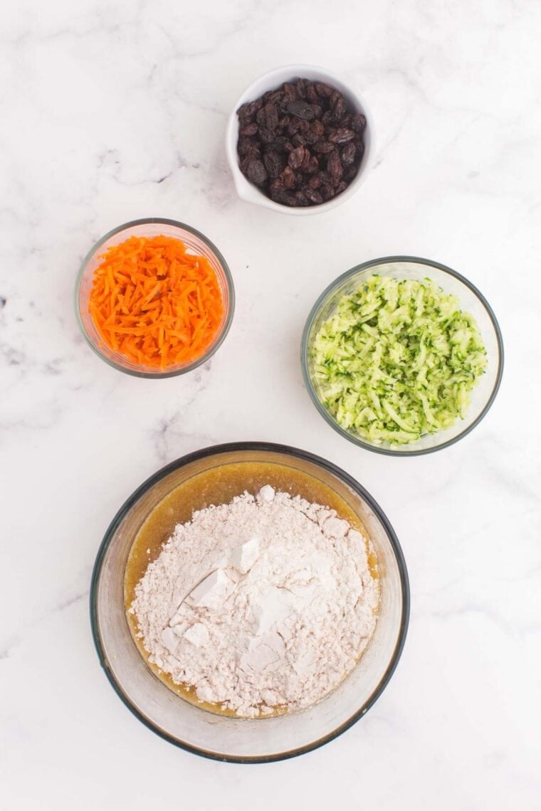 Dry ingredients added to the bowl of wet ingredients plus bowls of shredded carrots and zucchini, and raisins.