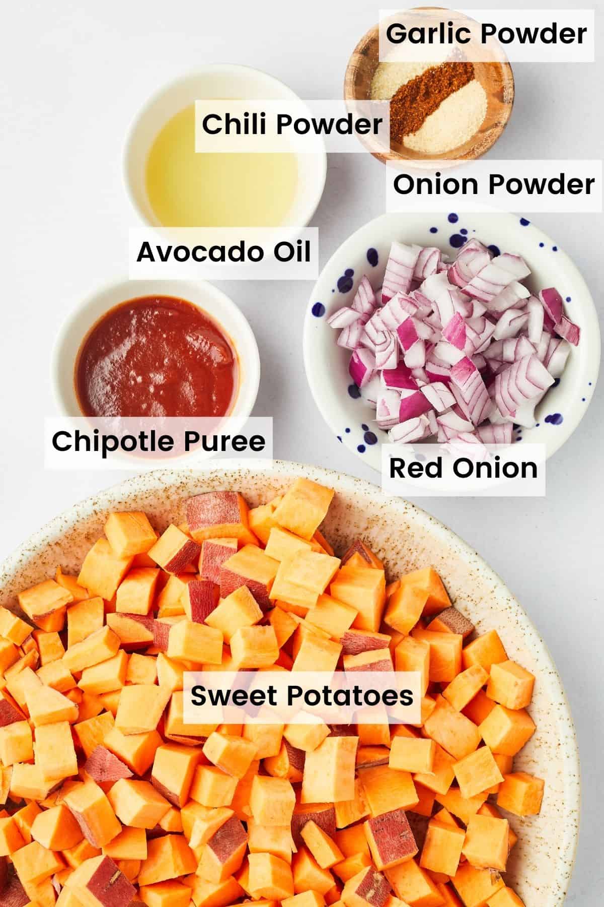 Ingredients for spicy air fryer sweet potato cubes are shown: sweet potatoes, red onion,, chipotles, onion powder, garlic powder, chile powder, avocado oil.