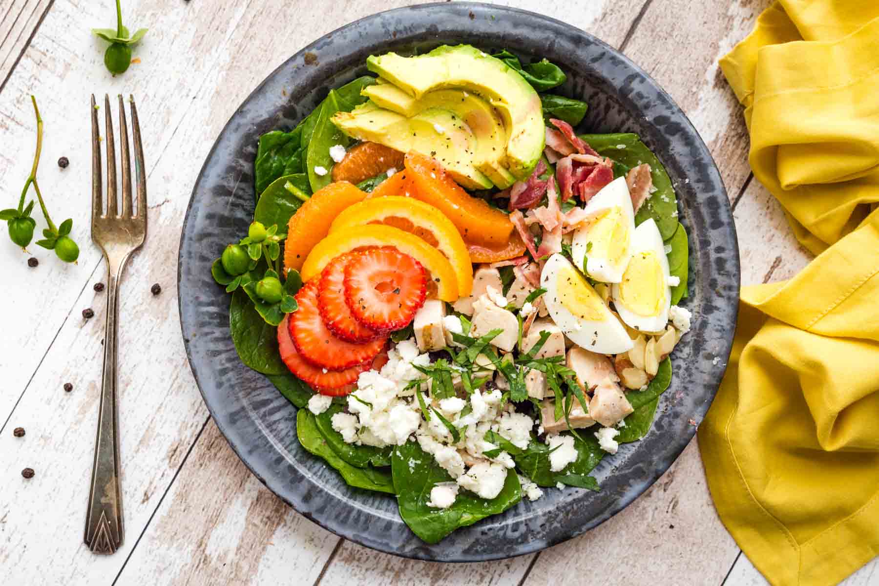 A bowl of strawberry spinach salad shows sliced avocado, strawberries, goat cheese, and hard boiled eggs.