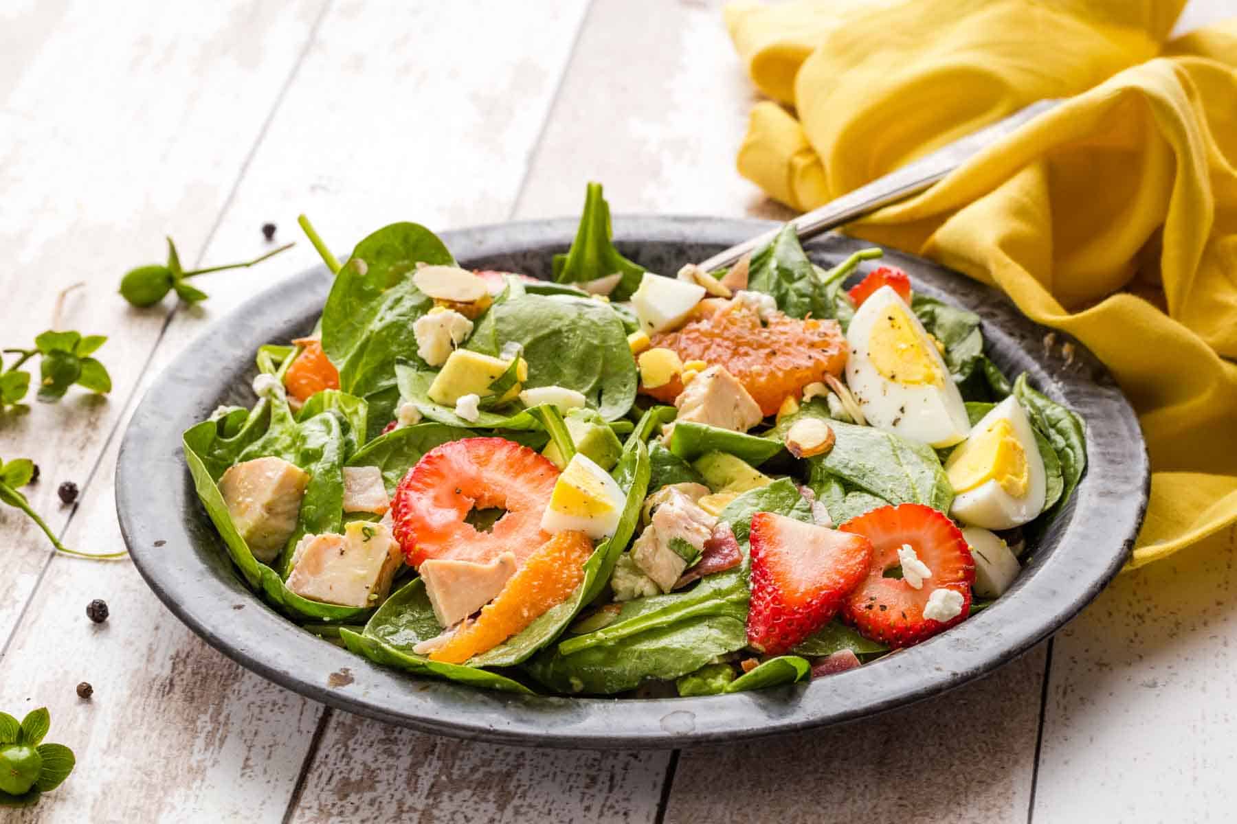 Slices of hard boiled eggs and strawberries along with spinach and goat cheese are shown close up in a bowl.