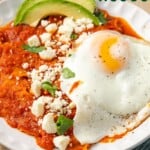 Tortilla chips cooked in a red sauce topped with a fried egg, cotija cheese, and avocado slices with text overlay that says "Chilaquiles Rojos".