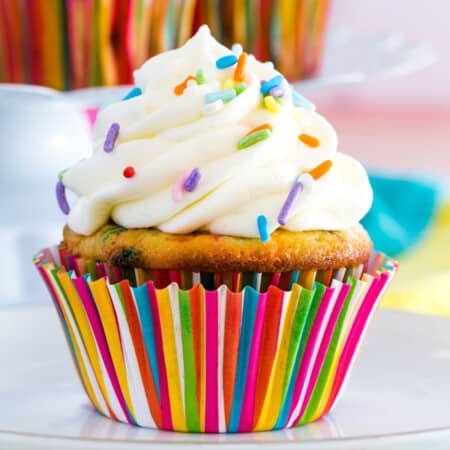 A cupcake in a colorful striped wrapper topped with a swirl of vanilla frosting and rainbow sprinkles.