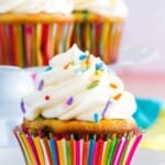 A cupcake in a colorful striped wrapper topped with piped vanilla frosting and rainbow sprinkles with text overlay that says "Gluten Free Funfetti Cupcakes".