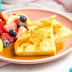 Two wedges of baked German pancakes on a pink plate with syrup and a side of fruit.