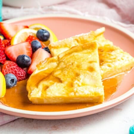 Two wedges of baked German pancakes on a pink plate with syrup and a side of fruit.