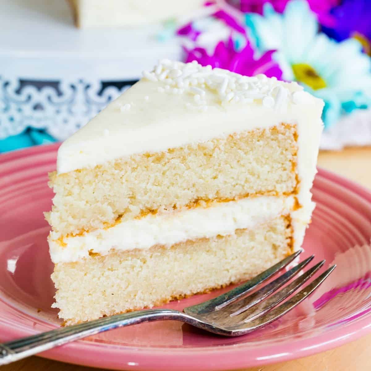A slice of white layer cake with vanilla frosting on a pink plate with a fork.