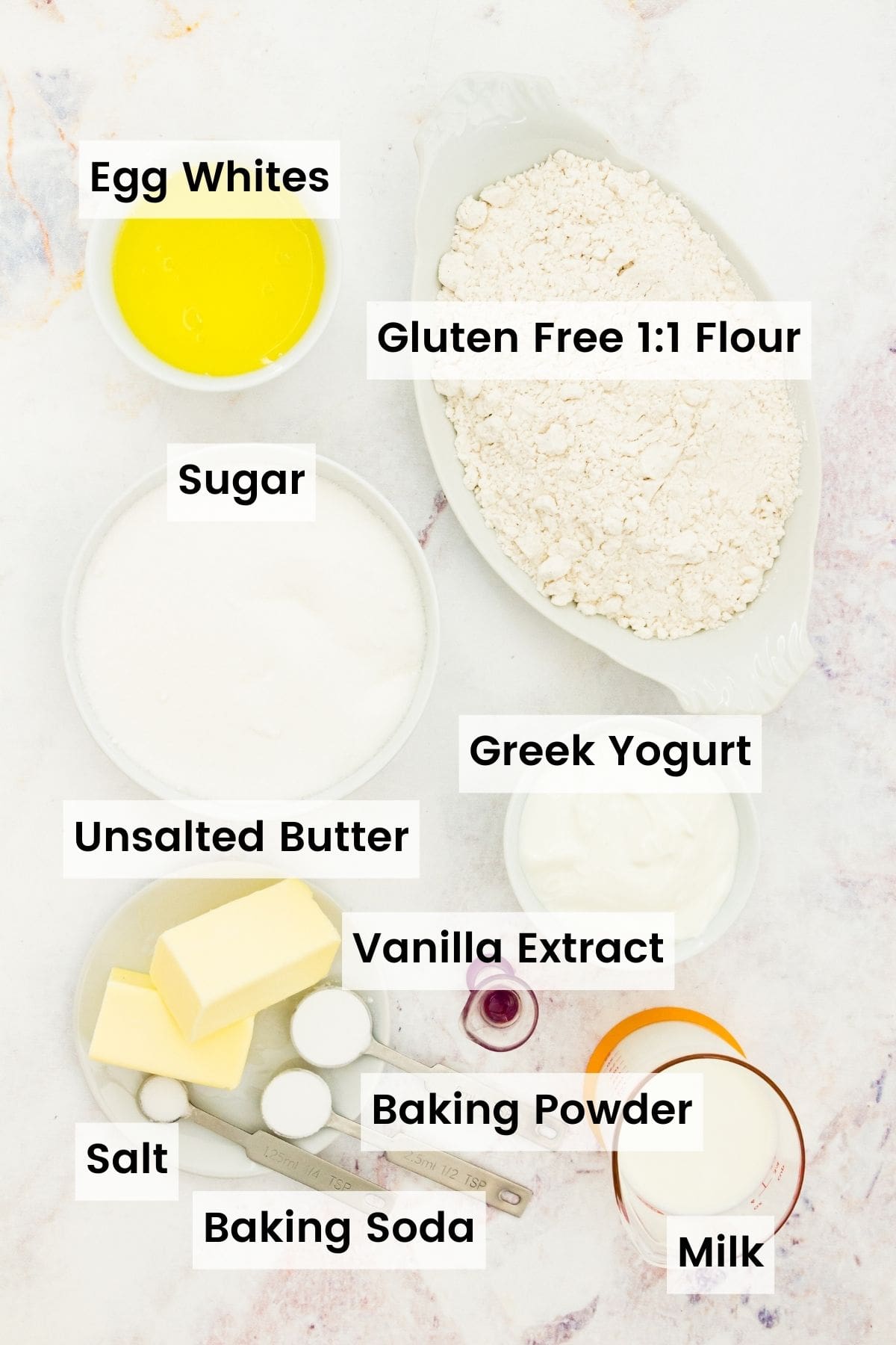 The ingredients for gluten free white cake are shown with text labels: gluten-free flour, vanilla extract, butter, egg whites, sugar, Greek yogurt, baking soda, baking powder, salt, milk.