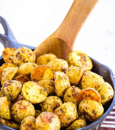 A wooden serving spoon stirs a cast iron skillet of pesto roasted potatoes.