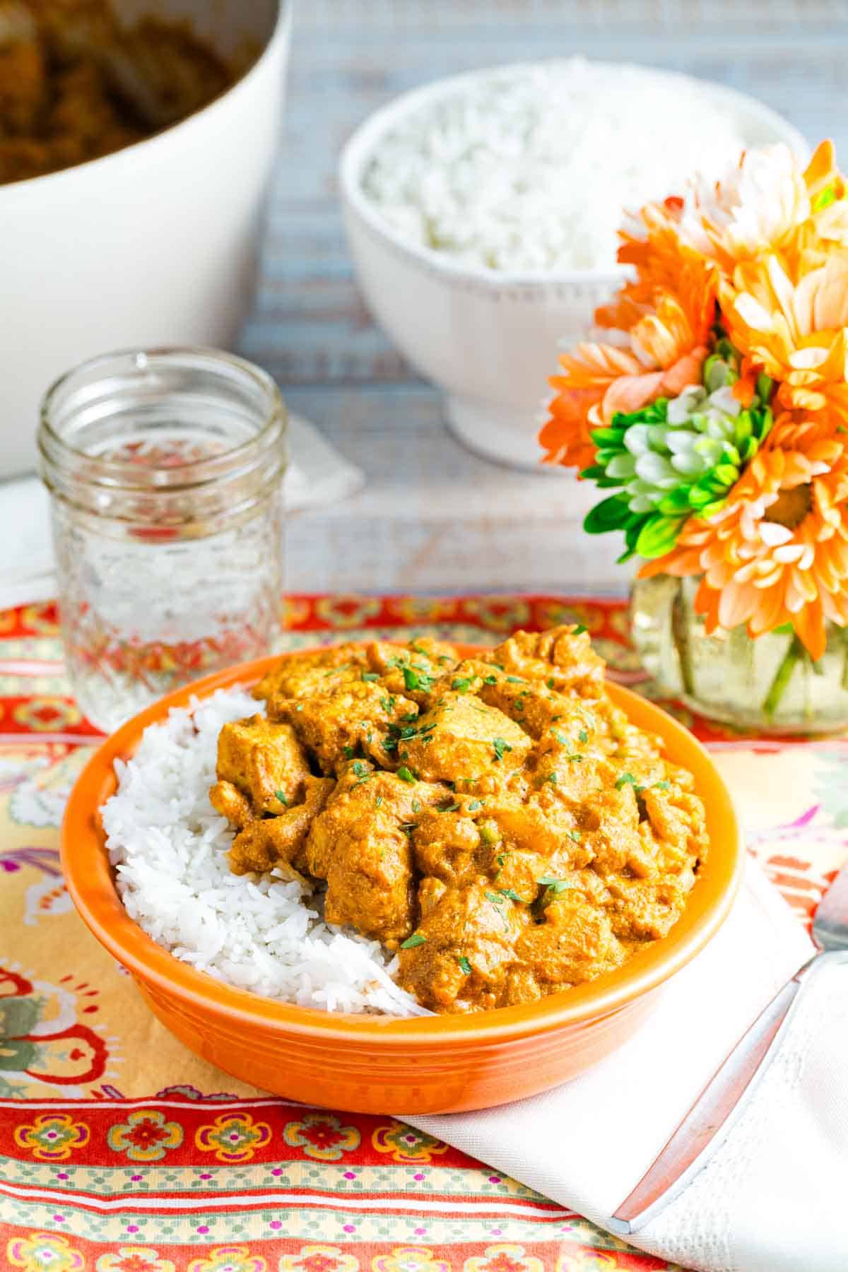 A serving of quick chicken curry and white rice is served in an orange bowl on a table with a napkin, glass of water, and flowers.