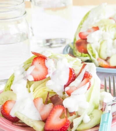 An iceberg wedge on a plate topped with strawberries, cucumbers, bacon, and blue cheese dressing.