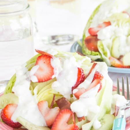 An iceberg wedge on a plate topped with strawberries, cucumbers, bacon, and blue cheese dressing.