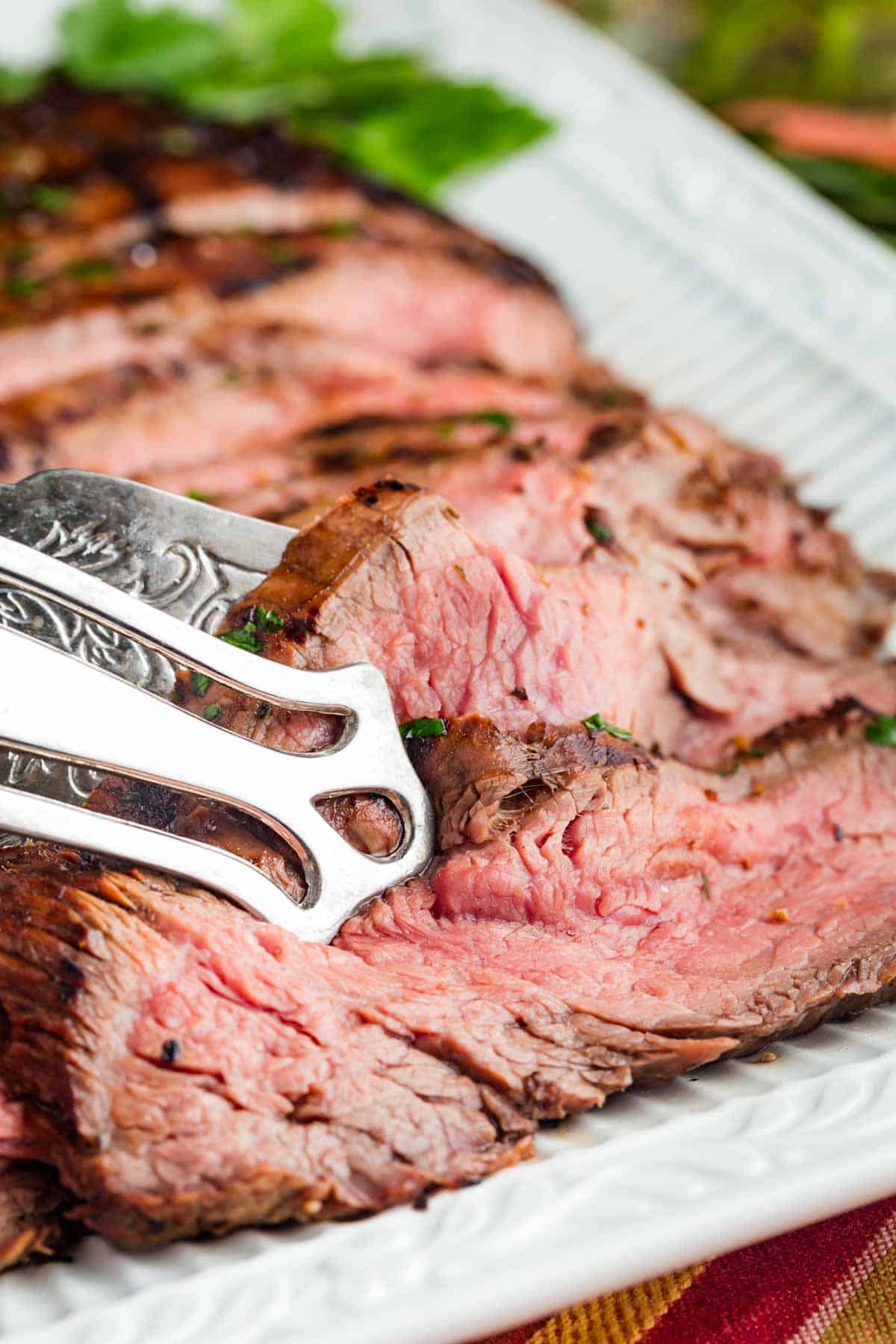 A serving tongs grabs a piece of grilled flank steak.