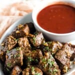 Seared pieces of steak garnished with parsley in a white dish with a bowl of red dipping sauce with text overlay that says "Garlic Butter Steak Bites".