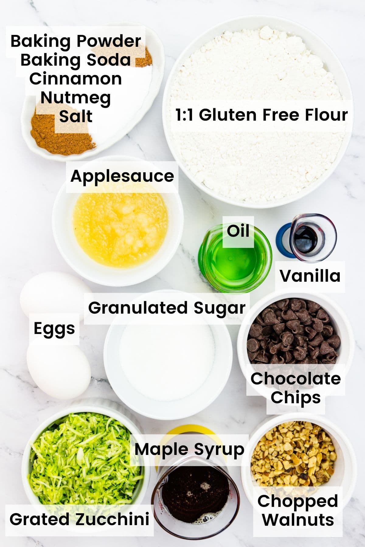 Ingredients for gluten free zucchini muffins are shown portioned out and labelled: zucchini, sugar, eggs, nutmeg, salt, baking powder, gluten free flour, maple syrup, chocolate chips, walnuts, vanilla.