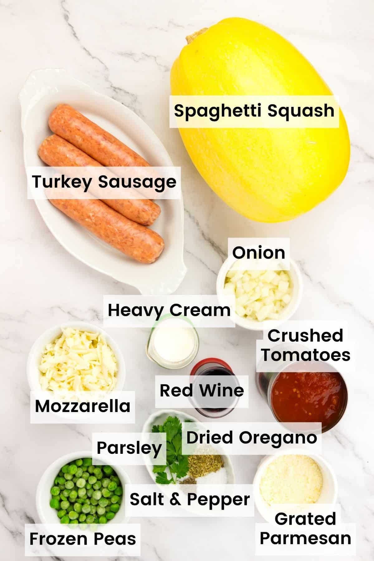 Ingredients for baked spaghetti squash on a countertop with text labels including: spaghetti squash, turkey sausage, onion, heavy cream, crushed tomatoes, red wine, mozzarella, dried oregano, parsley, salt and pepper, frozen peas, and grated parmesan.