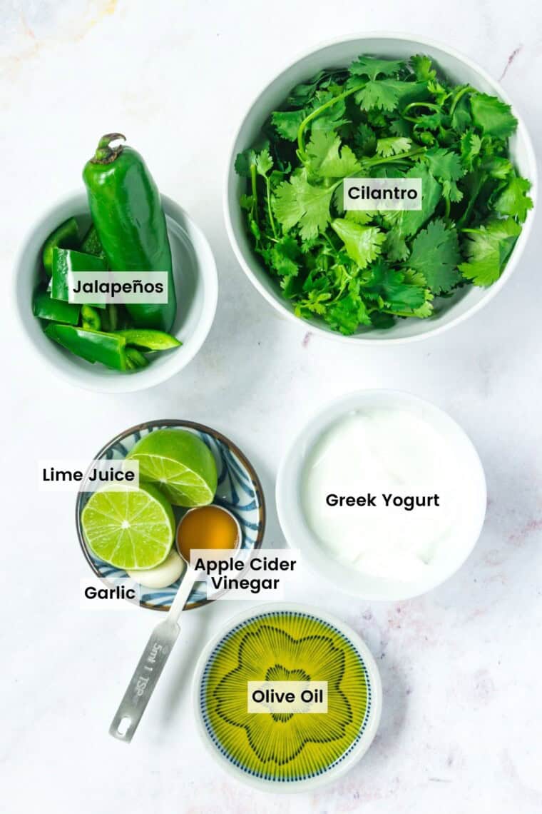 Ingredients to make Aji sauce in bowls on a marble countertop with text labels including cilantro, jalapenos, lime juice, garlic, apple cider vinegar, Greek yogurt, and olive oil.